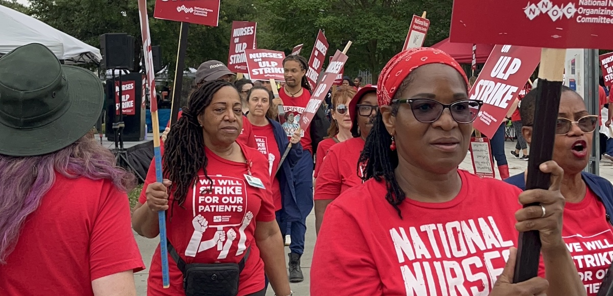 A group of nurses in red T-shirts with signs walk towards the camera.