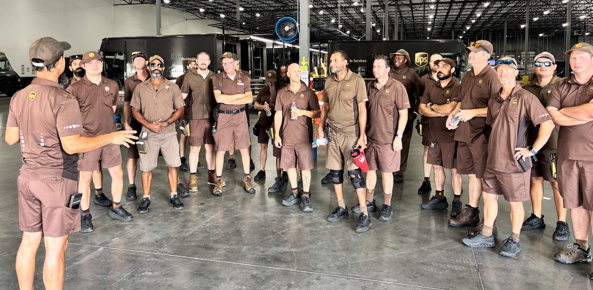Fifteen workers in brown UPS uniforms stand in a semicircle while another uniformed worker speaks to them