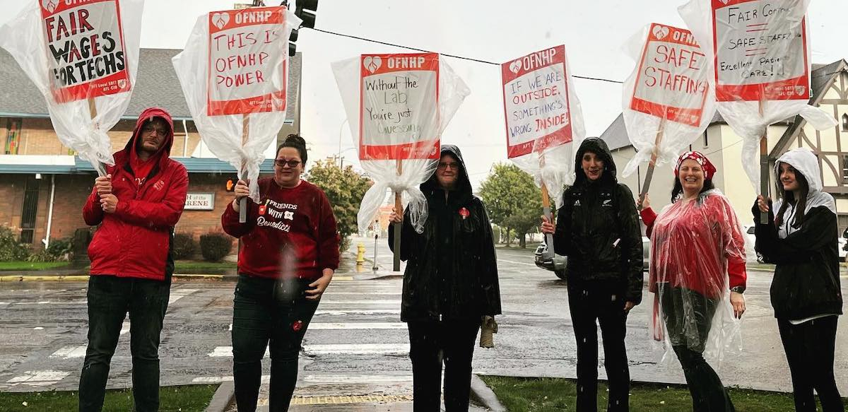 A group of six people stand facing the camera in the rain with signs covered with clear plastic bags. One says, ‘Without the Lab, You’re Just Guessing.’