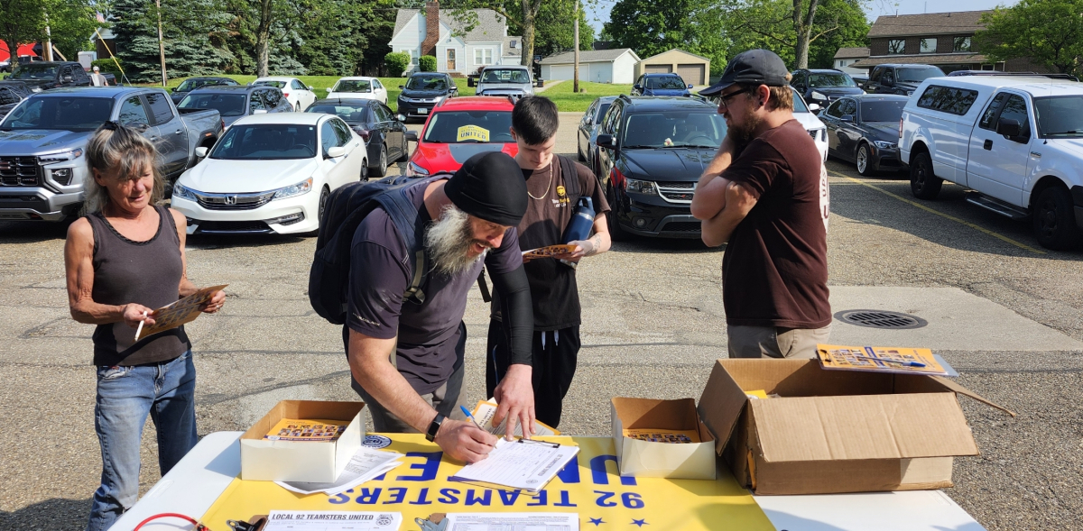 A group of Teamsters in a parking lot stand around a table with petitions and leaflets on it. One is filling out a sign-up sheet.