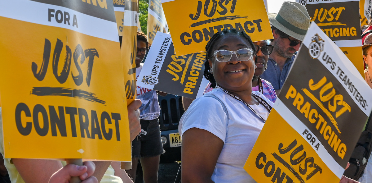 A Black woman in sunglasses smiles at the camera. She is holding a yellow and brown picket sign that says "UPS Teamsters, Just Practicing for a Just Contract." She is surrounded by matching signs, presumably held by other people who are mostly not visible in the shot.
