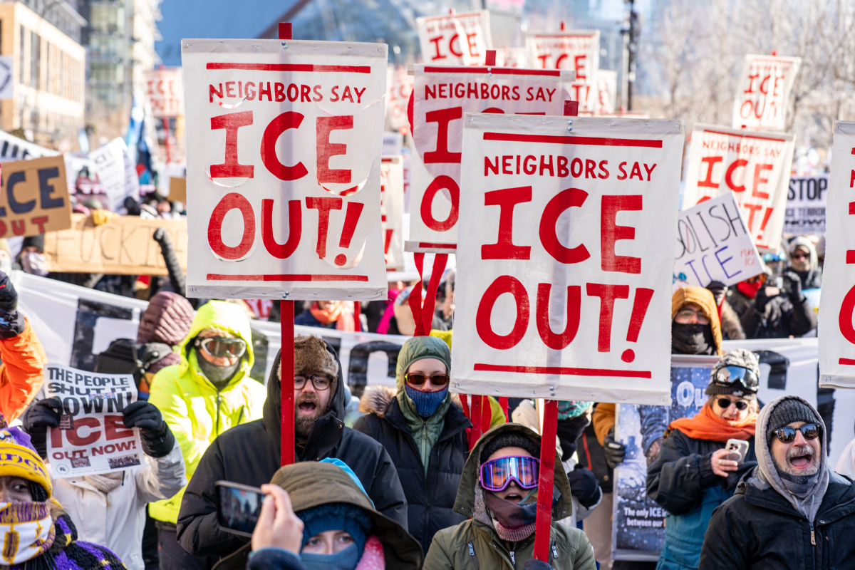View of a section of a giant crowd marching, on a bright sunny day, bundled against extreme cold. Prominent picket signs that look screen-printed or stenciled say "Neighbors say ICE out!" in crisp red on white background.