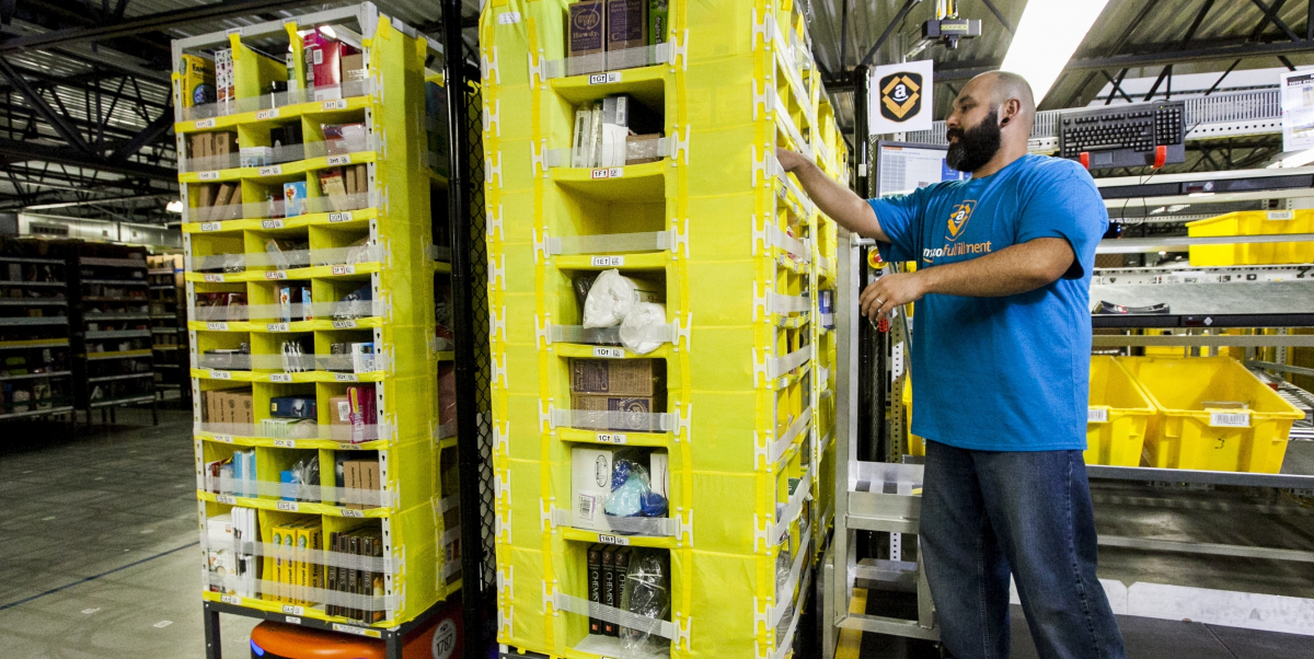 A worker in an Amazon warehouse picks items from a yellow shelf mounted on a robot.