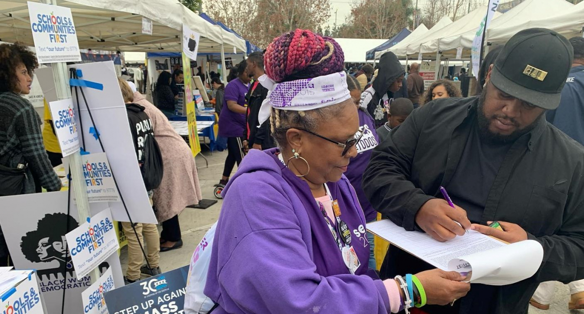 Outdoors at a public event, a woman in SEIU purple holds a clipboard while a man signs. The booth behind her is decked witht "SCHOOLS AND COMMUNITIES FIRST" signs and one that says "Step up against mass incarceration."