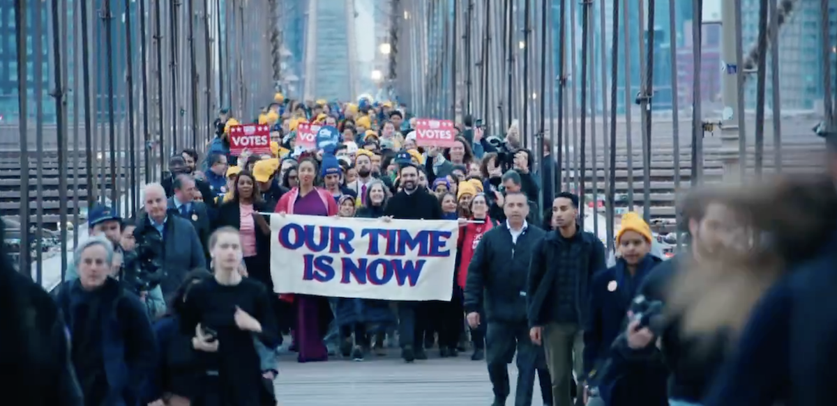 A dense and diverse crowd of people, some wearing yellow hats, marches across the Brooklyn Bridge in early-morning light. At the front are Zohran Mamdani and others carrying a banner that says in big purple letters "Our Time Is Now"