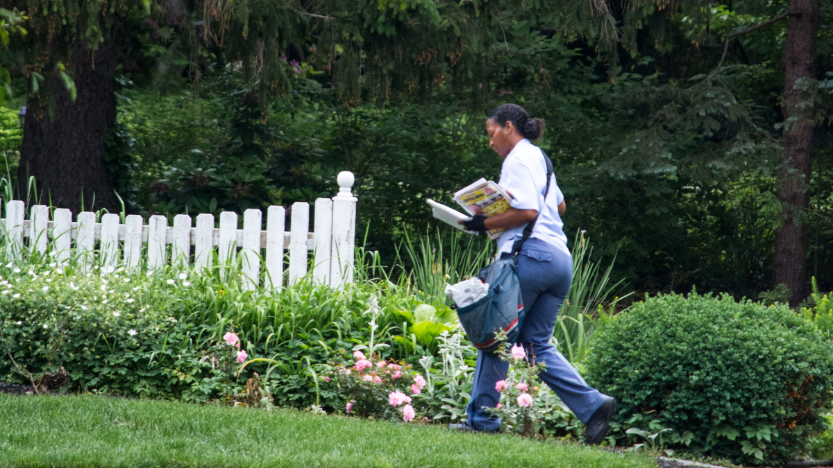 A Black woman letter carrier walks across a lawn, in front of a white picket fence, carrying the mail