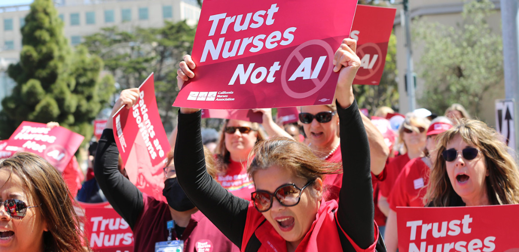 Women workers in red scrubs rally and chant, outdoors on a sunny day. They hold printed NNU signs. The woman most visible, in big sunglasses, holds a printed sign reading "Trust nurses, not A.I."