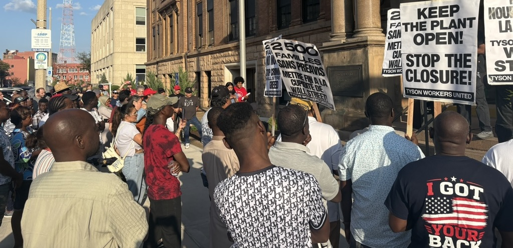 A group of workers, mostly Black, face an official-looking building with signs.