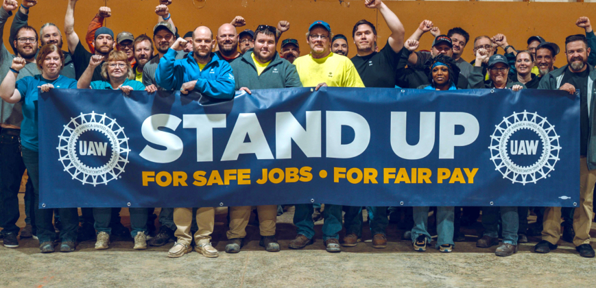 Thirty people, many with fists raised stand behind a banner that says “Stand Up, for safe jobs, for fair pay”