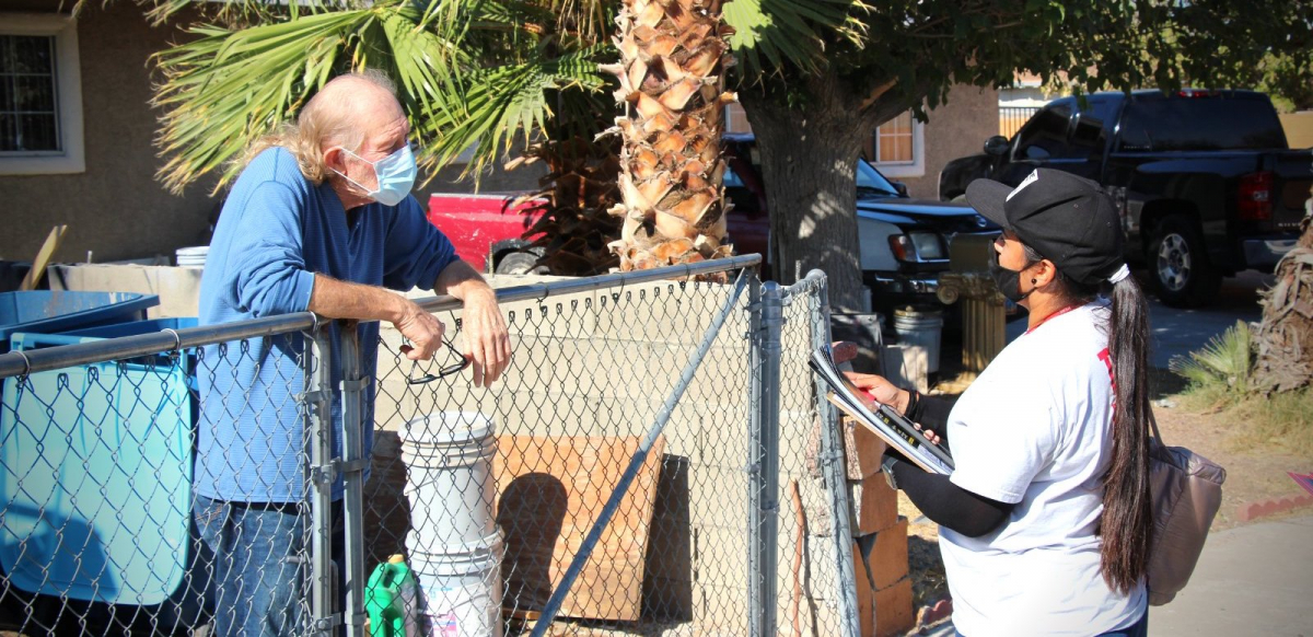 A Latina canvasser wearing a mask and holding a clipboard speaks to an older man leaning on his gate about the election