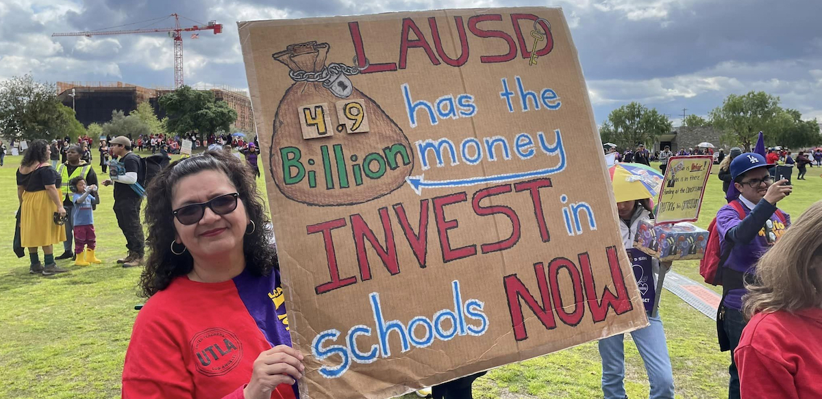 A woman in a red UTLA t-shirt holds a cardboard sign with red, blue, white and green lettering showing a big bag of money and saying “LAUSD has the money, 4.9 billion, invest in schools now.” She’s standing in a field with other demonstrators in the background.