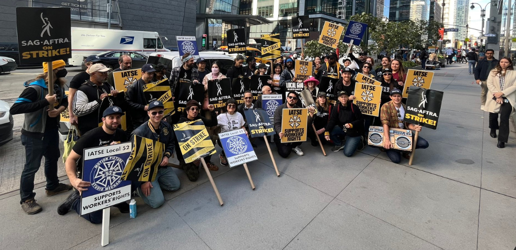 A large, upbeat crowd of picketers poses on a midtown Manhattan sidewalk, many kneeling. Some picket signs say "SAG-AFTRA on strike" and others say "IATSE supports workers' rights" or "IATSE Local 52 supporters workers' rights."