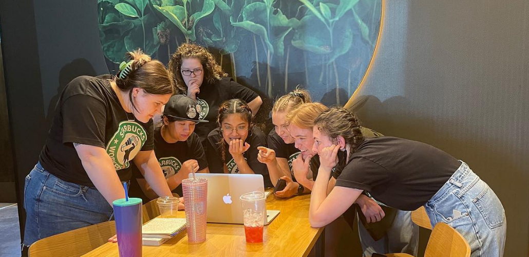 Young workers lean over a table, all peering intently at one laptop screen. They look excited and happy.