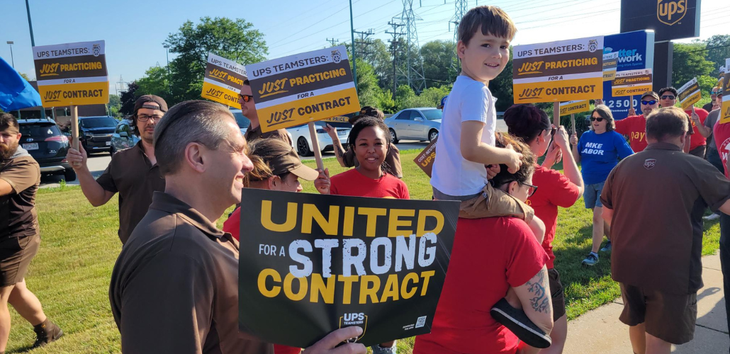 Workers picket with "United for a strong contract" and "Just practicing" teamsters signs. Some wear brown UPS driver uniforms. One little kid, riding on a parent's shoulders, turns to look happily directly into the camera.