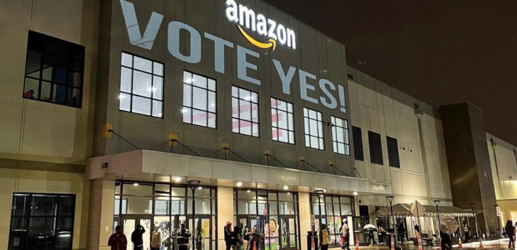Warehouse lit up against dark sky. Directly under the "Amazon" logo the message "Vote yes!" is projected on the wall. A few people are visible coming and going near the doors below.