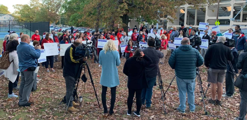 A crowd of red-shirted people stands outside Lawrence High School. One person is speaking to the group; others are shooting video of the event.