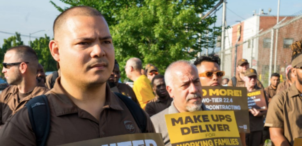 Crowd in UPS brown uniforms stands outdoors looking resolute, some holding signs. One sign visible reads "Make UPS Deliver!"