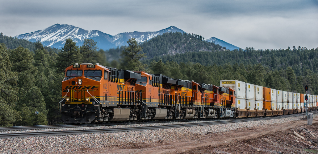 An orange BNSF train passes in front of a mountain and forested hills