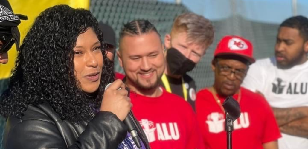 A woman speaks into a mic in foreground. Others wearing ALU T-shirts look on in background. The speaker and the listener nearest her are smiling. They are outdoors and it is sunny.