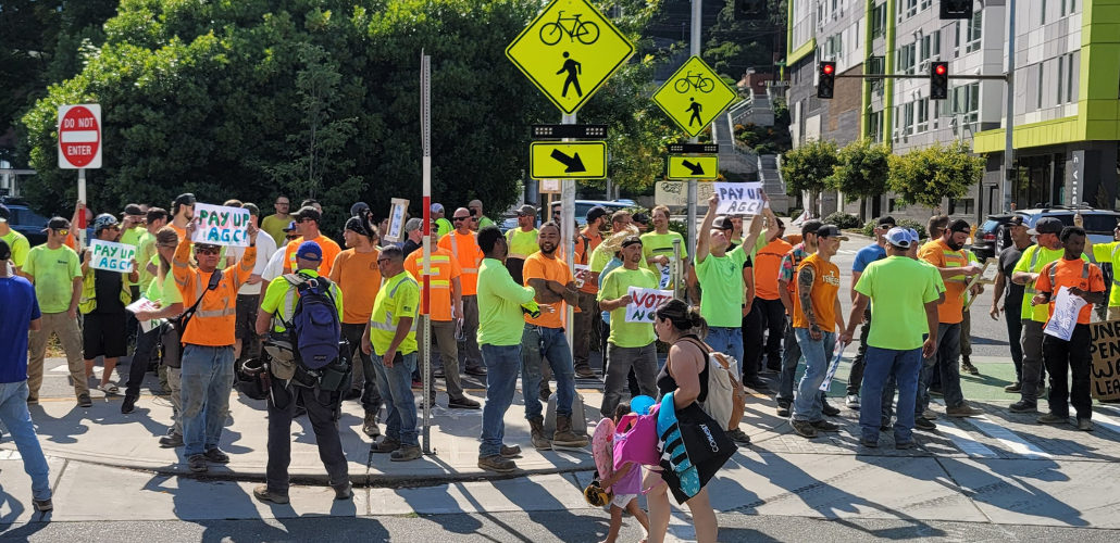 Big crowd of carpenters in high-vis neon orange and green shirts stands on a street corner. Some hold handmade signs reading "PAY UP AGC" and "VOTE NO."