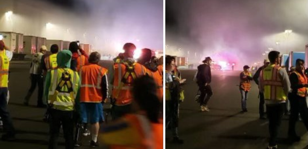 People in neon yellow and orange vests stand in front of clouds of purple smoke outside a warehouse at night.
