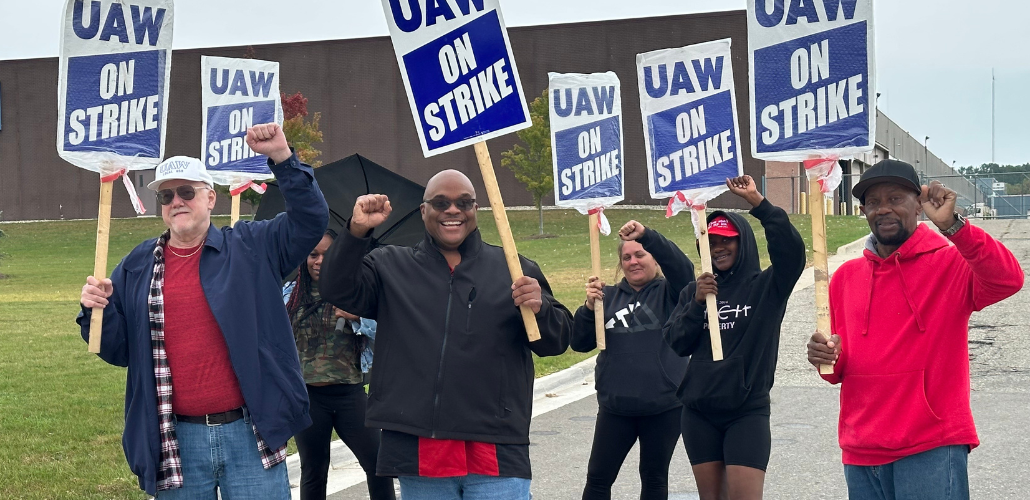 Six smiling workers carrying "UAW On Strike" picket signs raise their fists in the air outside a GM CCA facility. The workers are a mix of Black and white, men and women. Some of the picket signs are wrapped in plastic bags, and one worker carries an open umbrella.