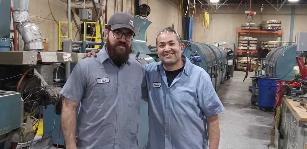 Two men in blue work shirts with nametags "Dan" and "Mohamed" sewn on stand side by side, arms around each other's shoulders, in a machine shop