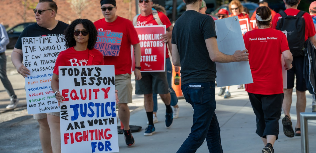 Strikers picket in red shirts. Most prominent is a woman with a sign: "Today's lesson: equity and justice are worth fighting for." Behind her is a man with a partially obscured sign listing "Part-time jobs I've worked so I can afford to be a teacher in Massachusetts" including Lyft driver, nightclub bouncer, Doordash, adjunct. 