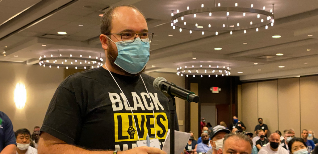 white man in mask at mic in hotel banquet room, wearing "Black Lives Matter" shirt, speaks as other people look on