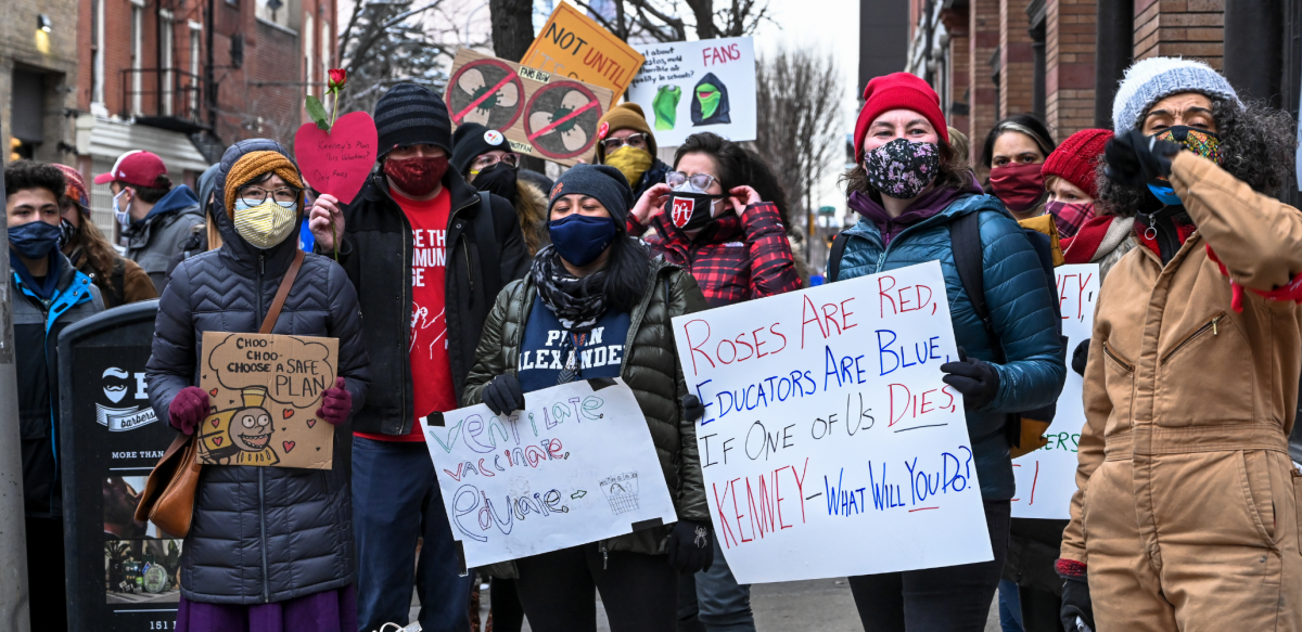 Crowd in coats and masks. One hand-drawn sign reads "Roses are red, educators are blue, if one of us dies, Kenney, what will you do?" Another shows a train and reads "Choo-choo-choose a safe plan." Another says "ventilate, vaccinate, educate." Another shows an image of a fan with a "no" line across it.