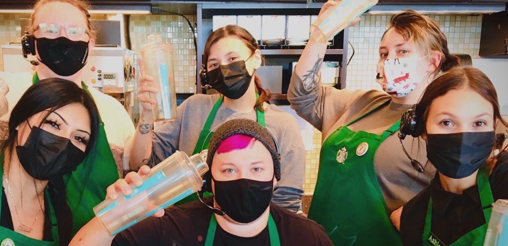 Six young workers in a Starbucks, wearing the trademark green aprons, look cheerfully or defiantly at the camera. Three are holding cups aloft.