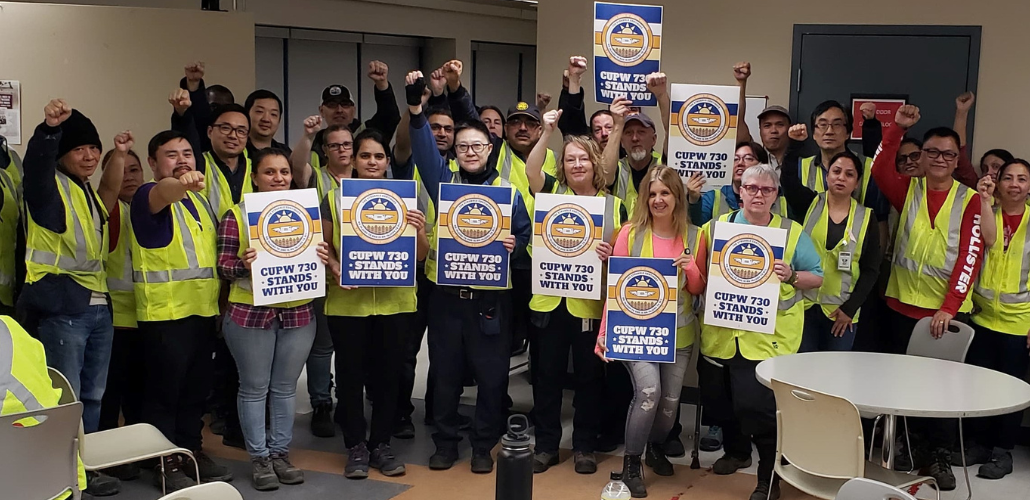 A group of smiling people in yellow vests stands inside a cafeteria. Several hold signs "CUPW Local 730 stands with you." Many have their fists in the air.