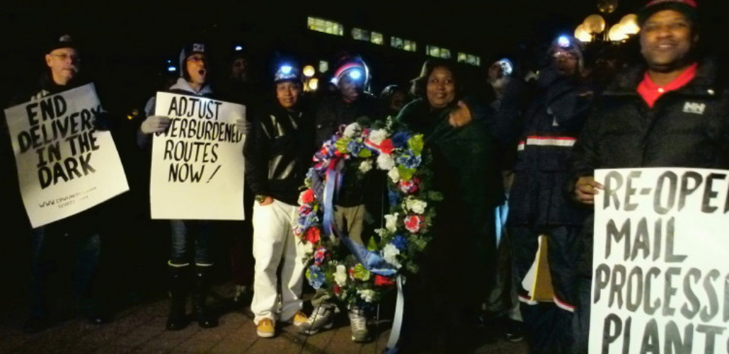 People in postal gear stand in the dark holding hand-lettered signs: "END DELIVERY IN THE DARK," "ADJUST OVERBURDENED ROUTES NOW," REOPEN MAIN PROCESSING PLANTS" and a memorial wreath.