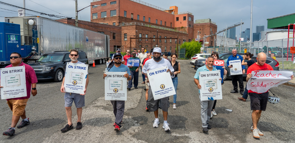 Workers walk forward outdoors carring signs reading "ON STRIKE: United Metro Energy Company: Teamsters Local 553." One person has a hand-lettered sign: "Teacher solidarity" that is flopping in the wind. Behind them is a brick industrial building. 