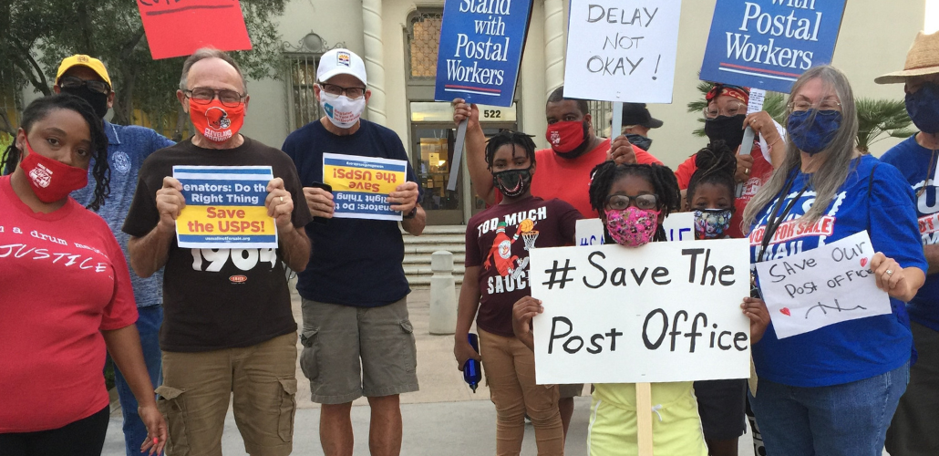 Masked people (Black and white, men and women) holding signs. Most prominent is a handwritten sign: "#Save the Post Office." Preprinted signs say "Stand with Postal Workers" and "Senators, do the right thing! Save USPS"