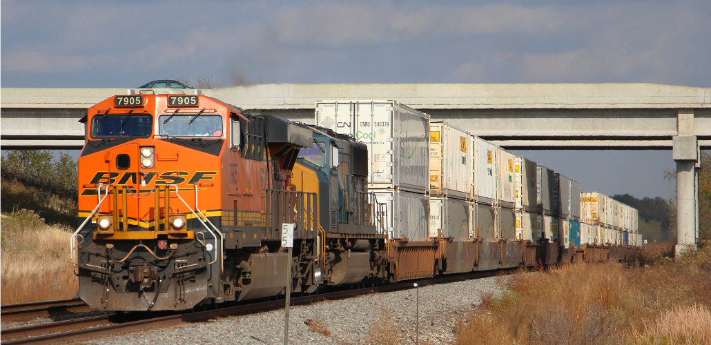 A freight train passes along a track under an overpass. The orange engine is labeled BNSF. It is pulling many white freight cars.