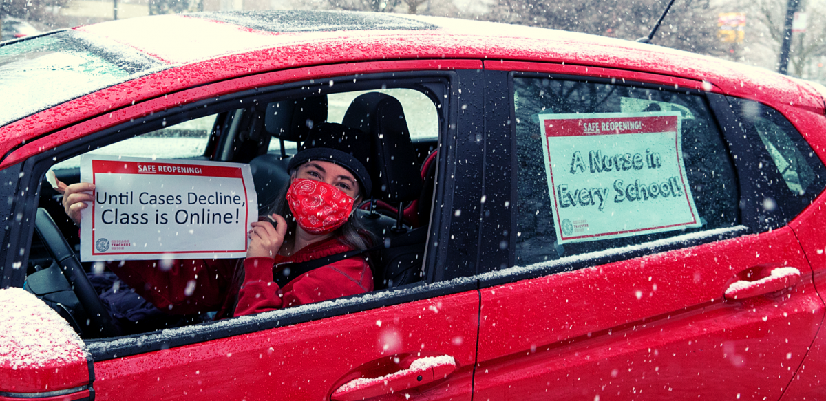 Person in driver's seat of red car, wearing red mask, holds sign: "Safe reopening! Until cases decline, class is online!" Taped to back window of car is another sign: "A nurse in every school!" It is snowing.