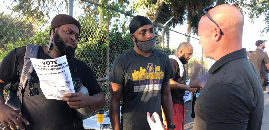 Sean O'Brien (right), a bald white man, speaks as two workers, Black men, listen. One worker is holding a Teamsters United flyer. Behind them is a chain-link fence and someone in a UPS delivery uniform.