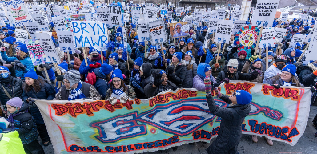 Huge, shouting crowd of people in blue hats, many with picket signs "On strike for smaller class sizes." One prominent handmade sign says "Bryn Mawr." A big banner at front of crowd says, in artsy spray-painted style, "District refuses to pay for ESPs, our children's education heroes!" The S in ESPs is the Superman logo. 