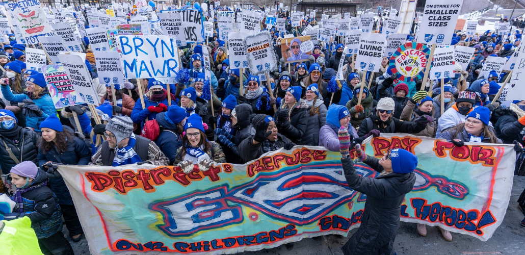 Huge, shouting crowd of people in blue hats, many with picket signs "On strike for smaller class sizes." One prominent handmade sign says "Bryn Mawr." A big banner at front of crowd says, in artsy spray-painted style, "District refuses to pay for ESPs, our children's education heroes!" The S in ESPs is the Superman logo.