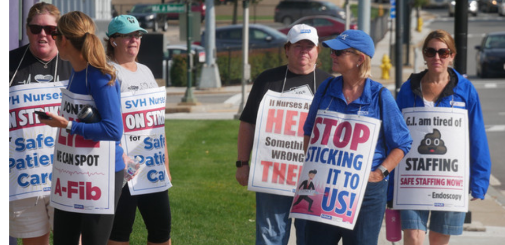 Several nurses picket outside wearing sign. One sign says "Stop sticking it to us!" with a drawing of a person in a business suit puncturing the letters with a giant syringe and needle. Another says "G.I. am tired of [poop emoji] staffing. SAFE STAFFING NOW. -Endoscopy." Other signs are partially obscured but appear to say "Don't lie, we can spot A-Fib," "If nurses are out here there's something wrong in there," and "SVH nurses on strike for safe patient care."