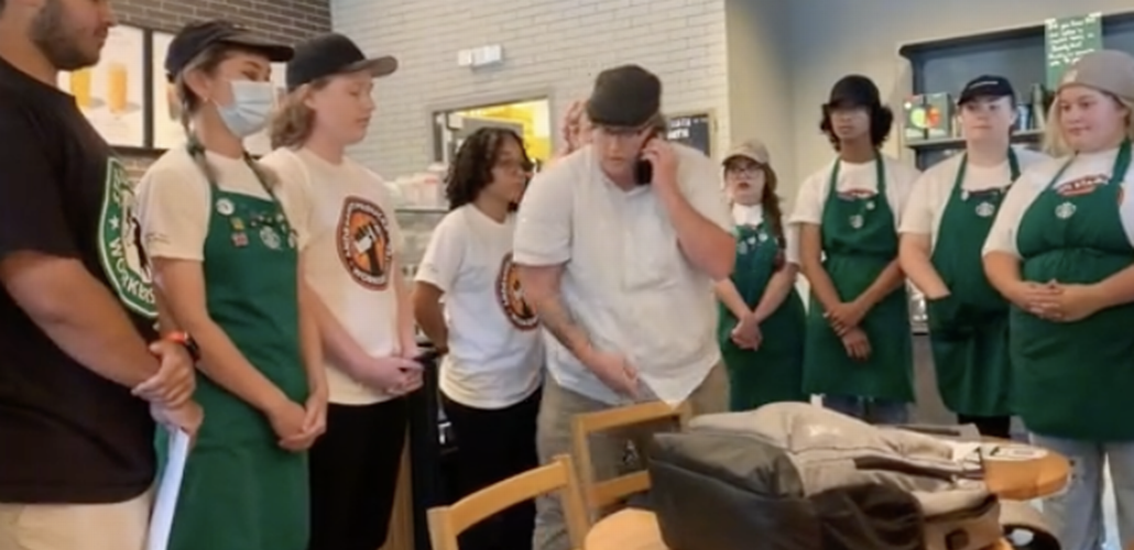 Young workers in Starbucks aprons and SBWU logo shirts stand in a semicircle, all in calm postures mostly with hands clasped at rest in front of them. In the center is the manager in a white shirt, talking into a cell phone.