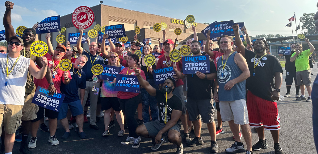 An energetic crowd of workers of various ages, genders, and races, Shawn Fain among them, poses in the sunshine outside a building marked Local 862. Many hold printed signs: "United for a strong contract," "Every job a good job," and the UAW logo. Many thrust their fists in the air.