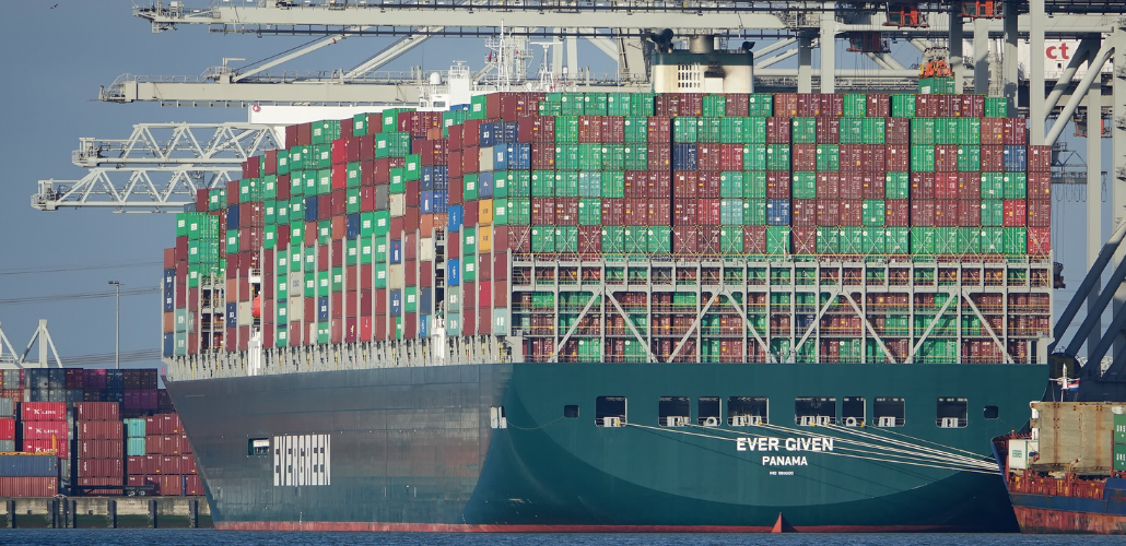 Huge ship loaded with containers, viewed from stern which says "Ever Given, Panama." In the background are port cranes.