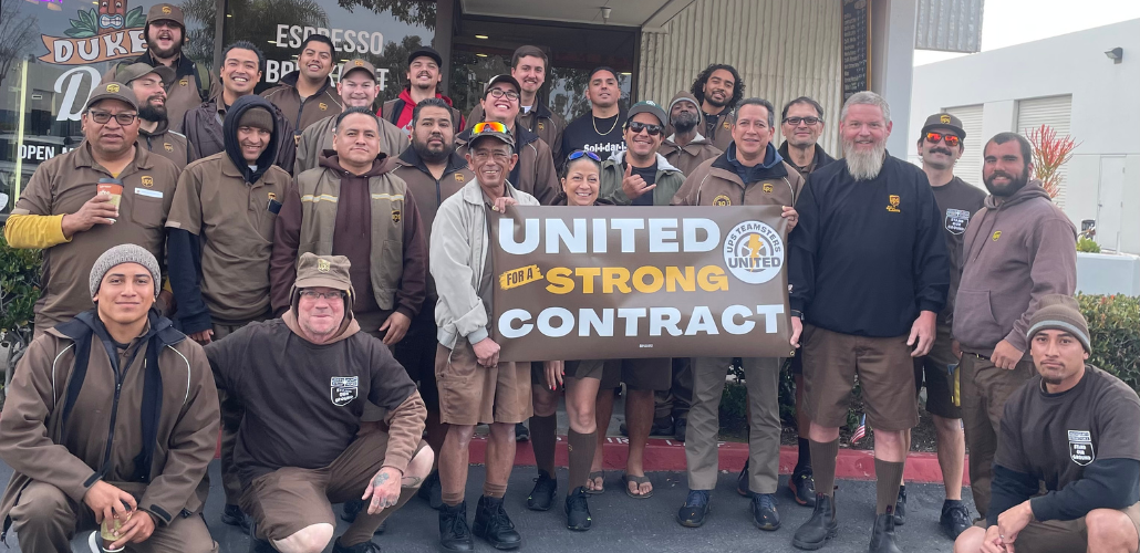 26 people (mostly men) in brown UPS uniforms pose outside Duke's coffee shop, smiling. Some hold a banner: "United for a strong contract" with the UPS Teamsters United logo.