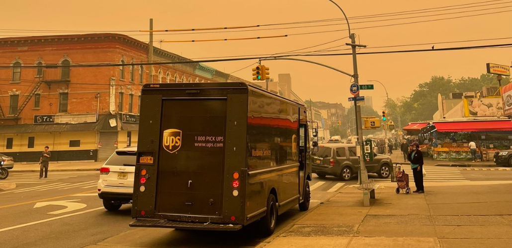 UPS truck viewed from behind on a Brooklyn street; visible are a few pedestrians and a fruit market. The sky and the whole scene is blanketed in a thick orange haze; buildings down the block are fuzzier because of the smoke.