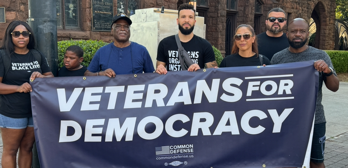 A group of veterans stand behind a blue banner that reads Veterans for Democracy.