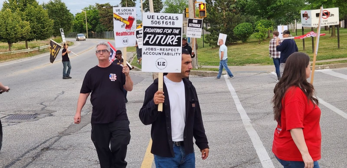 workers walk a circular picket in a driveway, with one visible sign saying “Fighting for our future: Jobs, respect, accountability.”