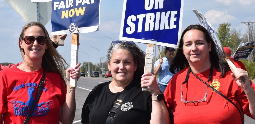 Three women smile at the camera with UAW on Strike signs.