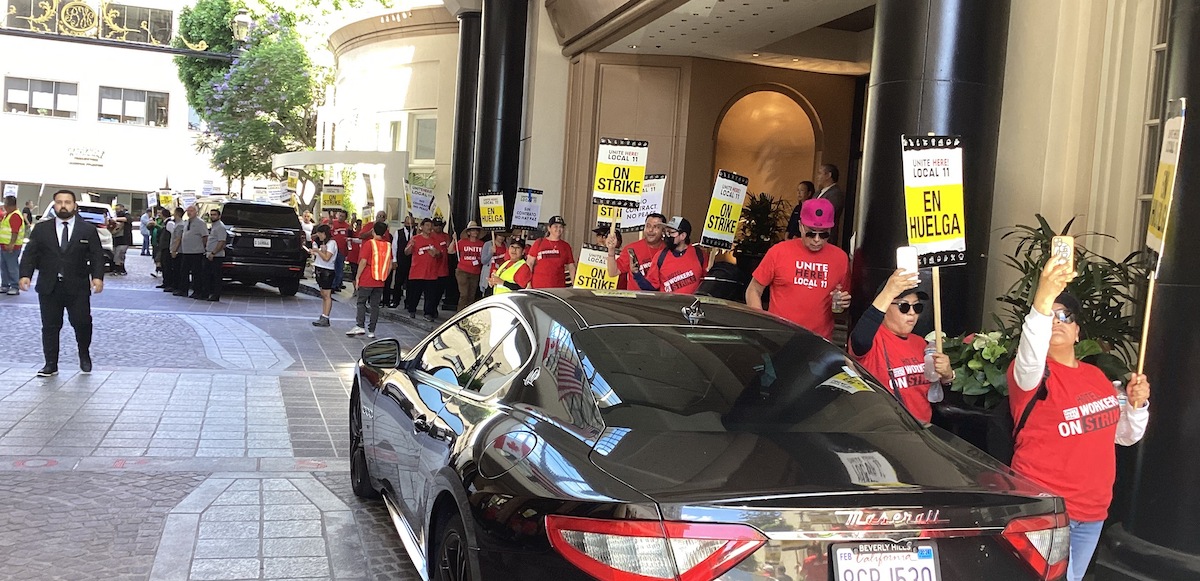 Hotel workers in red t-shirts march past the entrance of a fancy hotel carrying signs that say “Huelga” and “Strike.”.